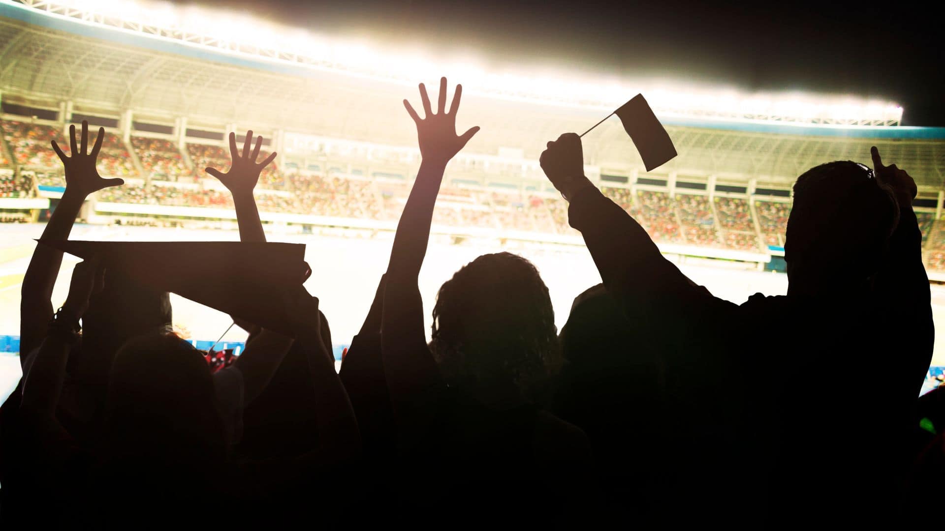 This image depicts a stadium crowd, with people silhouetted in the foreground. They have their hands raised in excitement, some holding flags, and appear to be cheering. The background shows a brightly lit stadium with a large number of spectators in the stands, giving the sense of a live sports or concert event. The overall atmosphere is energetic and celebratory.