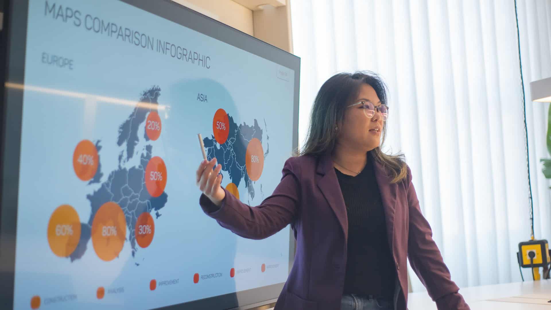 A woman in a purple blazer and glasses gives a presentation in front of a large screen displaying a world map infographic.